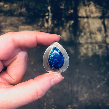 Vintage silver clip-on earrings with teardrop-shaped azurite malachite stones in shades of deep blue and green, held in a person's hand against a dark textured background.