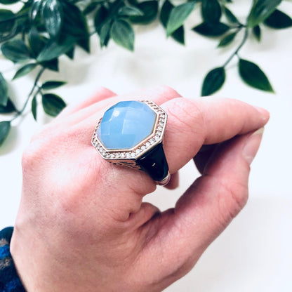 Vintage sterling silver cocktail ring with light blue stone surrounded by filigree design, shown on hand against leafy green background.