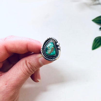 Vintage silver ring with a green turquoise stone set in an ornate setting, shown on a finger against a plain background with plant leaves.