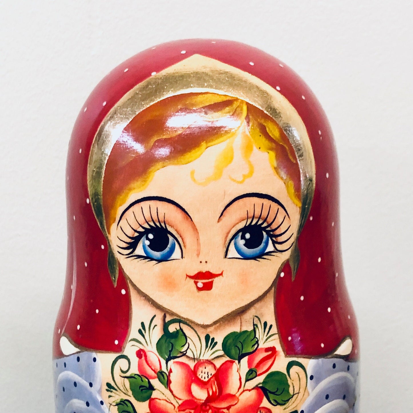 Close-up of a smiling blonde Matryoshka doll with blue eyes, rosy cheeks and wearing a red floral headscarf and blue polka dot dress, painted in a traditional Russian folk art style.