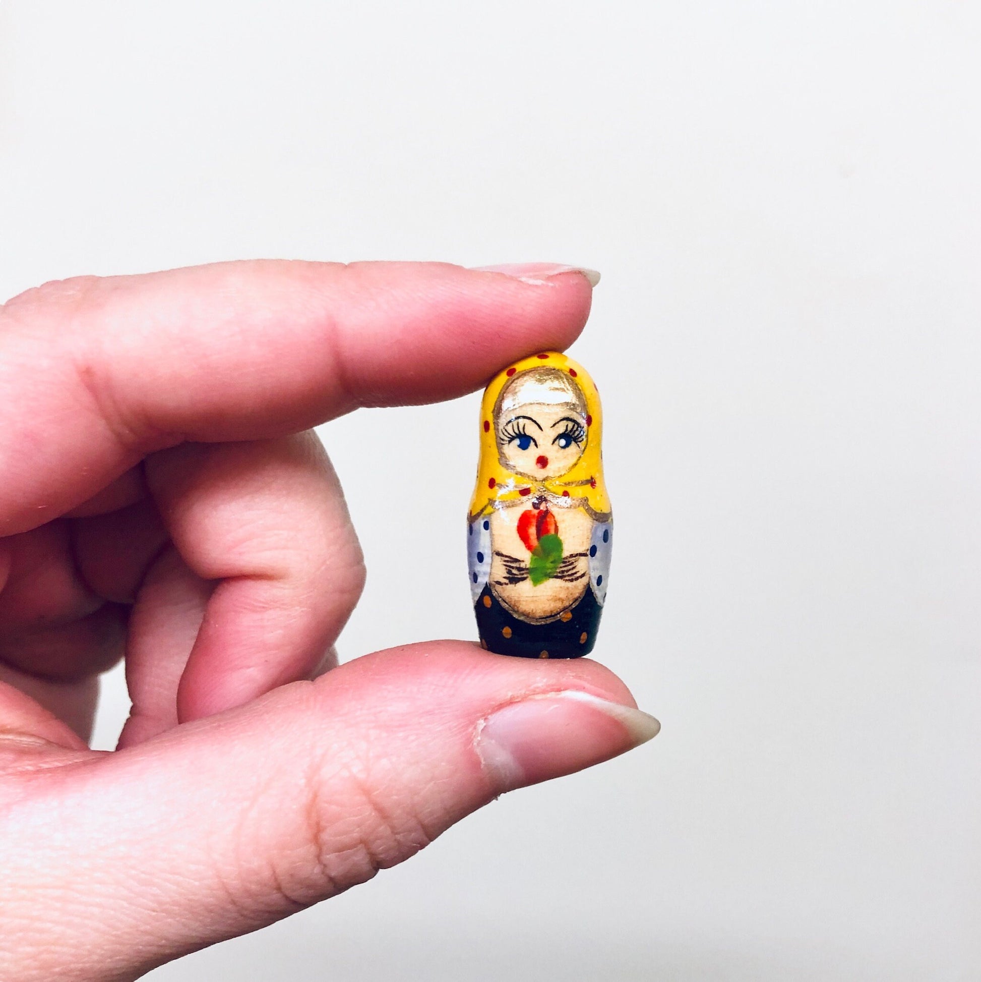 Hand holding a miniature Russian nesting doll painted with a blonde-haired female character wearing a yellow shawl and holding a rose against a white background.