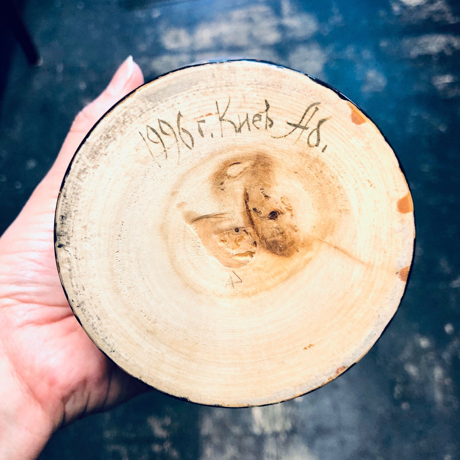 Close-up view of a tree stump cross-section showing rings and knots, with "1996 Hark AS" carved into the wood surface, held in a person's hand against a dark blue textured background.