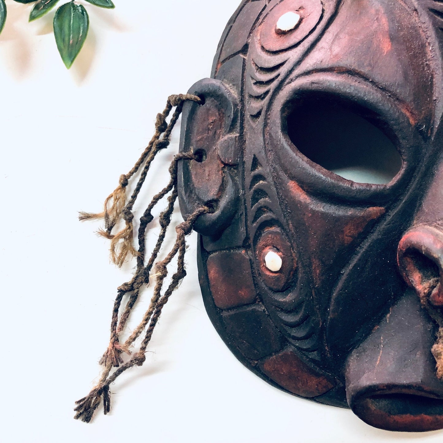 Vintage hand carved wooden tribal mask with intricate details and cutout design, hung on a white wall with leafy decor, unique wall art and home decor piece