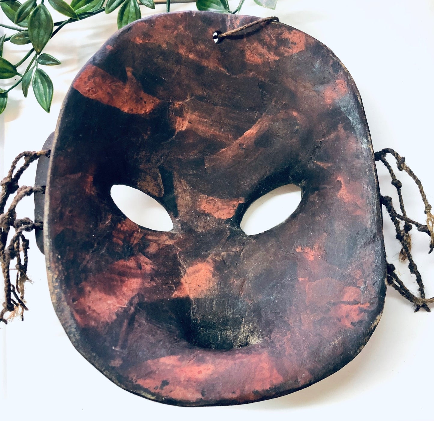 Vintage hand carved wooden mask with cut out eyes hanging on a white wall, surrounded by green leaves. The mask has a dark reddish-brown textured surface resembling an abstracted face.