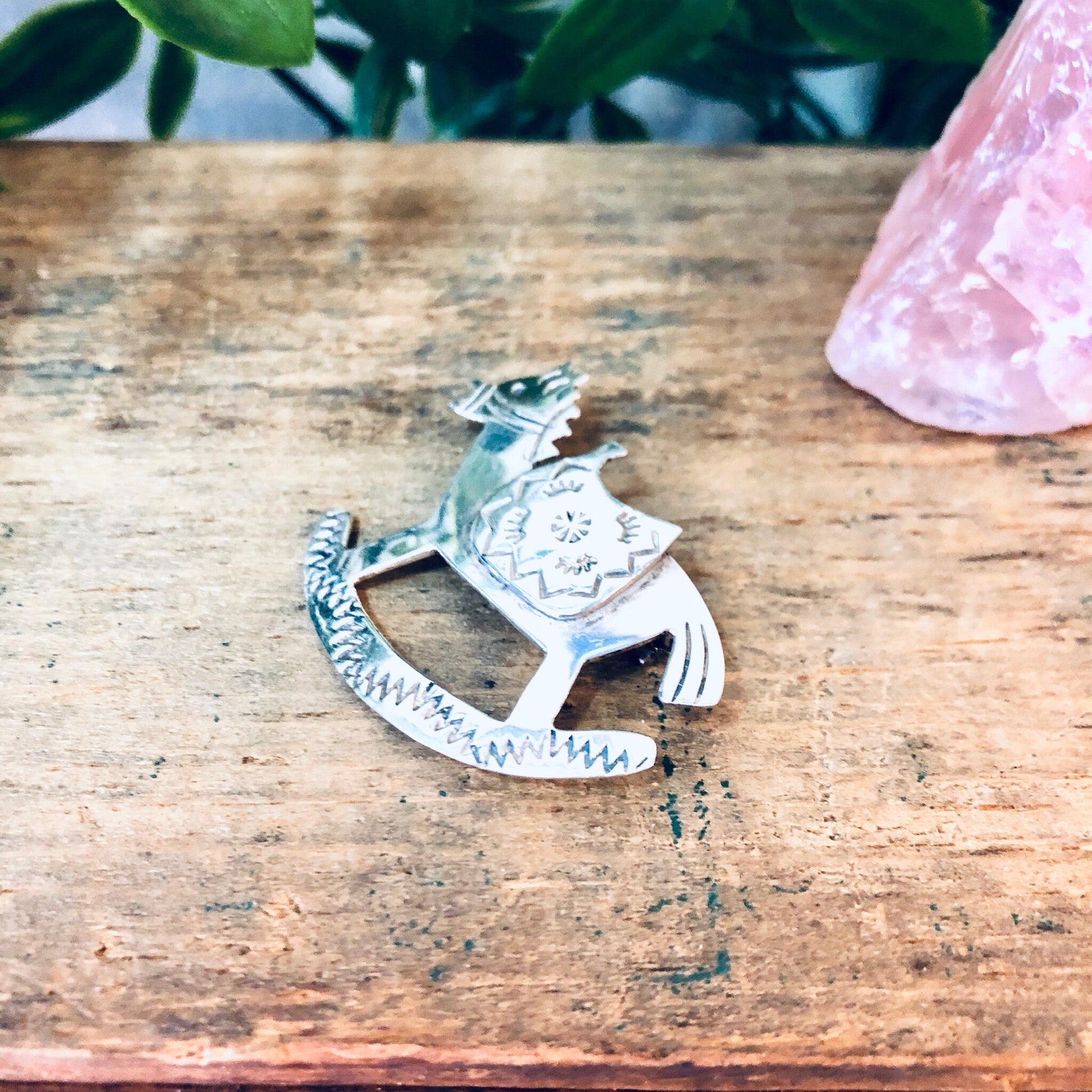 Vintage silver rocking horse brooch, made in Mexico, sitting on rustic wooden surface with pink crystal and plant in background.