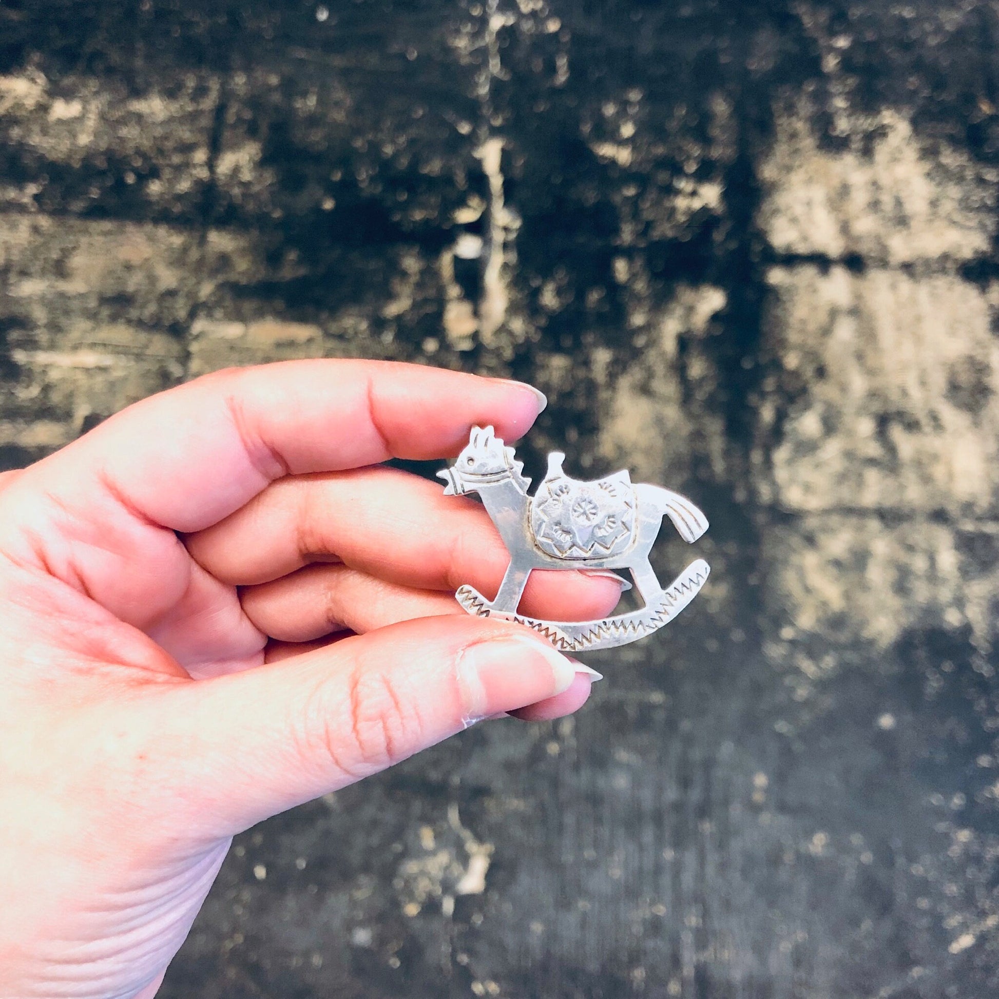 A hand holding a vintage silver rocking horse brooch against a weathered wooden background.