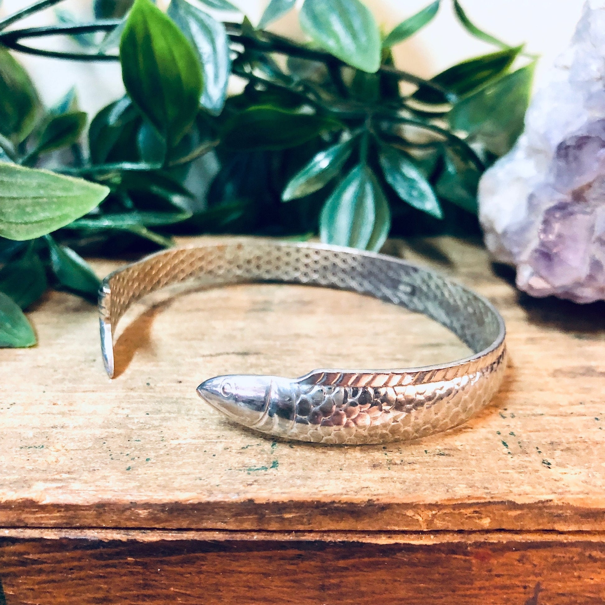 Vintage silver fish bangle bracelet on wooden surface with crystals and green leaves in background