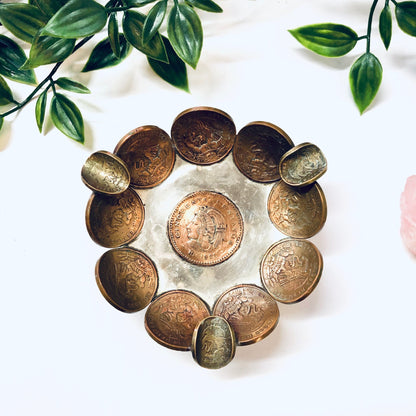 Vintage Mexican coin ashtray or catch-all dish made of copper centavos arranged in a circular pattern, surrounded by green leaves on a white marble surface.