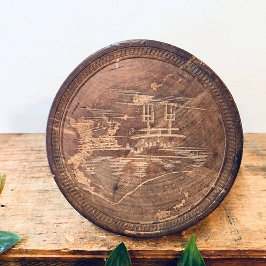 Antique wooden trinket box with intricate carving of house and trees on round lid, placed on weathered wood surface with leaves, unique rustic home decor or jewelry storage idea