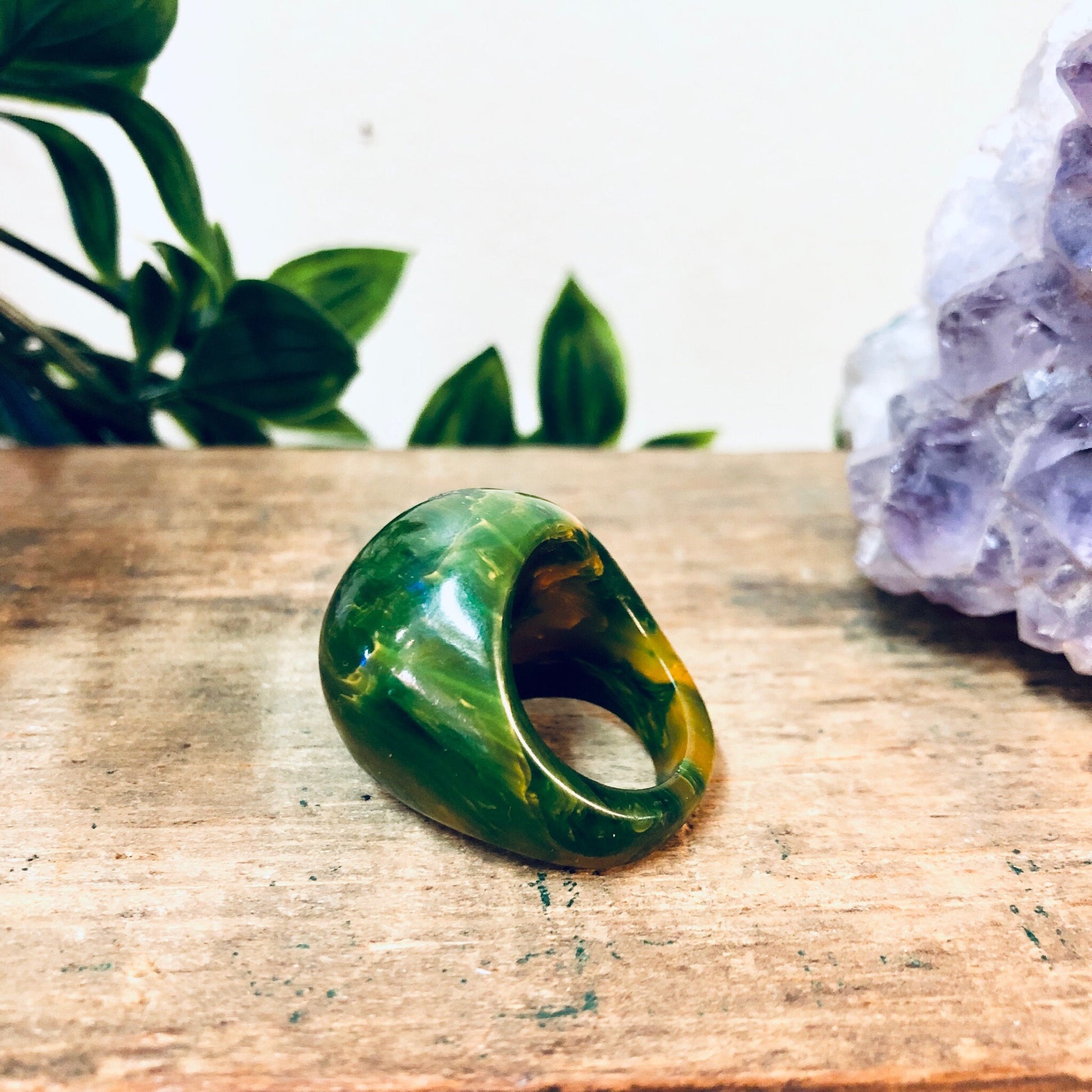 Green and yellow marbled plastic cocktail ring on wooden surface with amethyst crystal and plants in background, retro statement jewelry gift idea for Valentine's Day