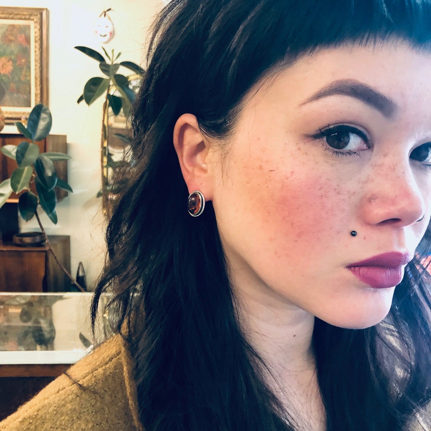 Close-up portrait of a young woman with long dark hair, freckles on her cheeks, and silver hoop earrings. She has a subtle smile and is looking slightly off-camera in a room with plants and framed artwork visible in the background.