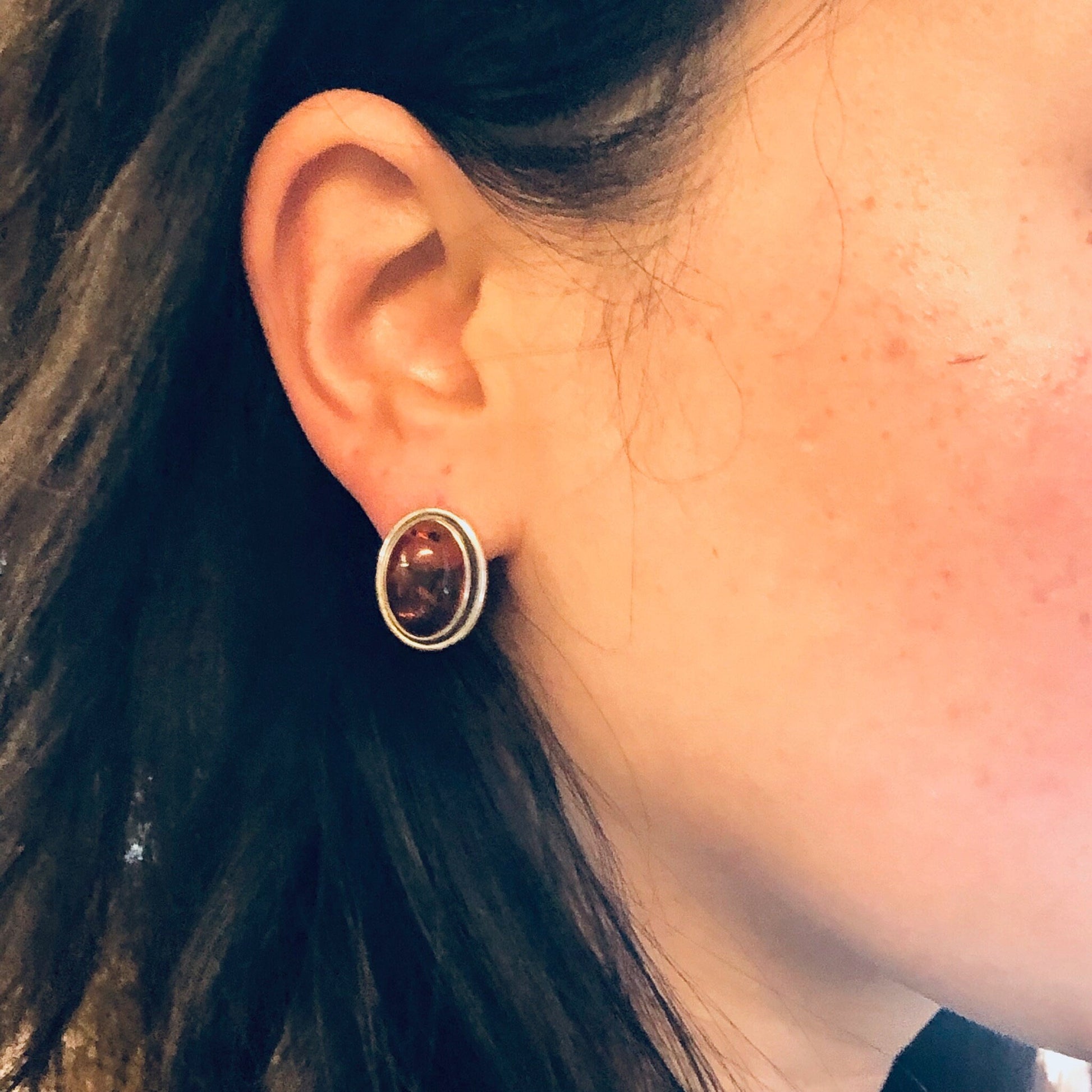 Closeup of a person's ear wearing an amber stud earring set in a silver frame, with strands of dark hair visible.