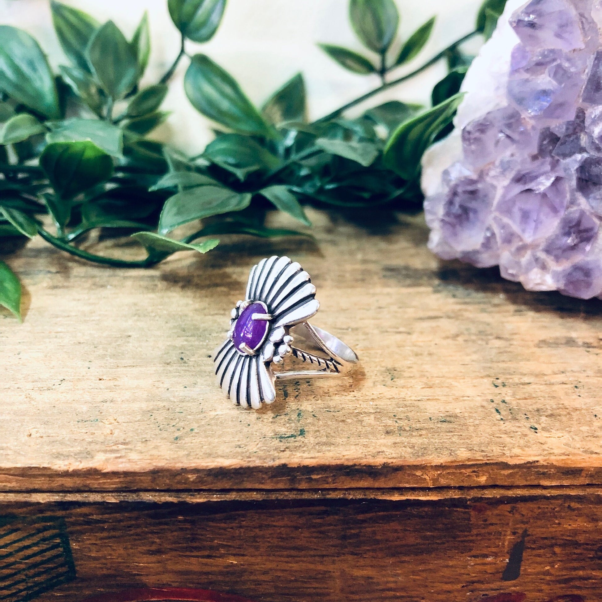 Vintage silver fan ring with purple gemstone on rustic wooden surface next to green leaves and purple amethyst crystal