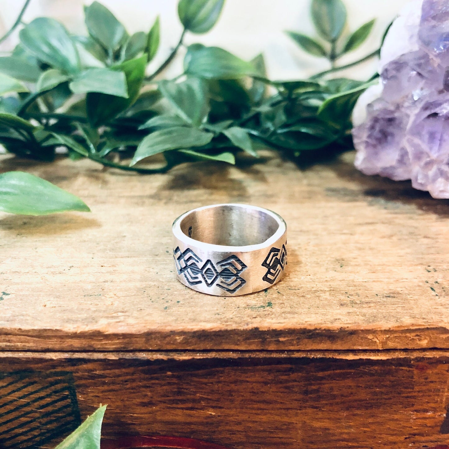 Vintage silver geometric tribal design thick band ring displayed on wooden surface with green plants in soft focus background