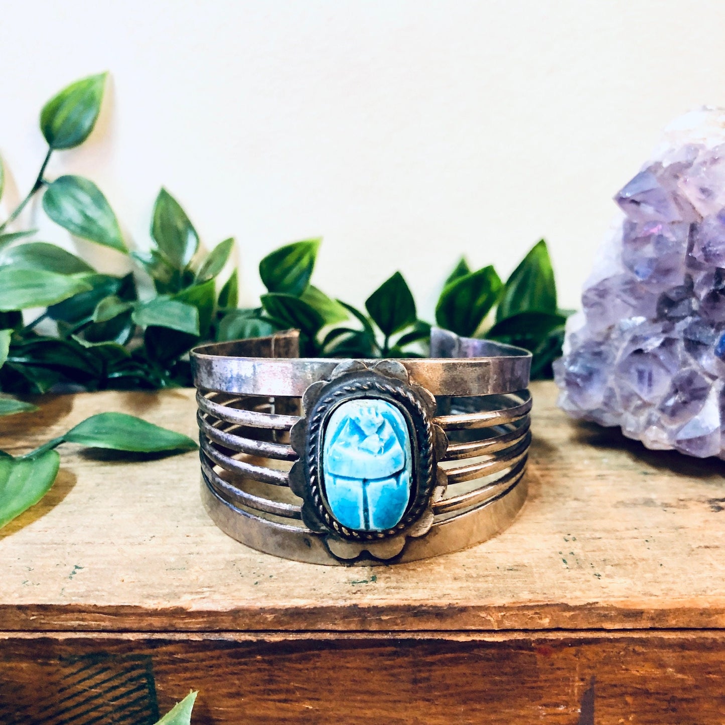 Vintage silver-toned cuff bracelet with blue scarab cabochon amidst green foliage and amethyst crystal, on rustic wooden surface.