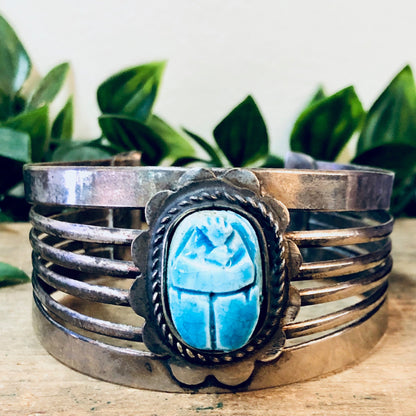 Vintage silver-toned cuff bracelet featuring a large blue scarab gemstone centerpiece, displayed on a wooden surface with green leaves in the background.