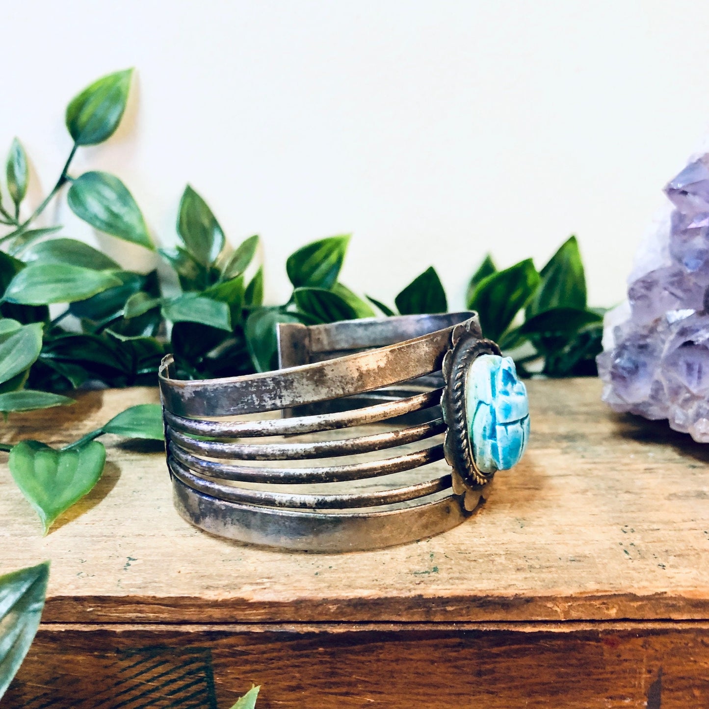 Vintage silver-toned cuff bracelet featuring a large blue scarab cabochon, displayed on a wooden surface with green foliage and an amethyst crystal in the background.