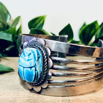 Vintage silver cuff bracelet with large blue scarab gemstone surrounded by detailed metalwork, placed on a wooden surface with green leaves in the background.