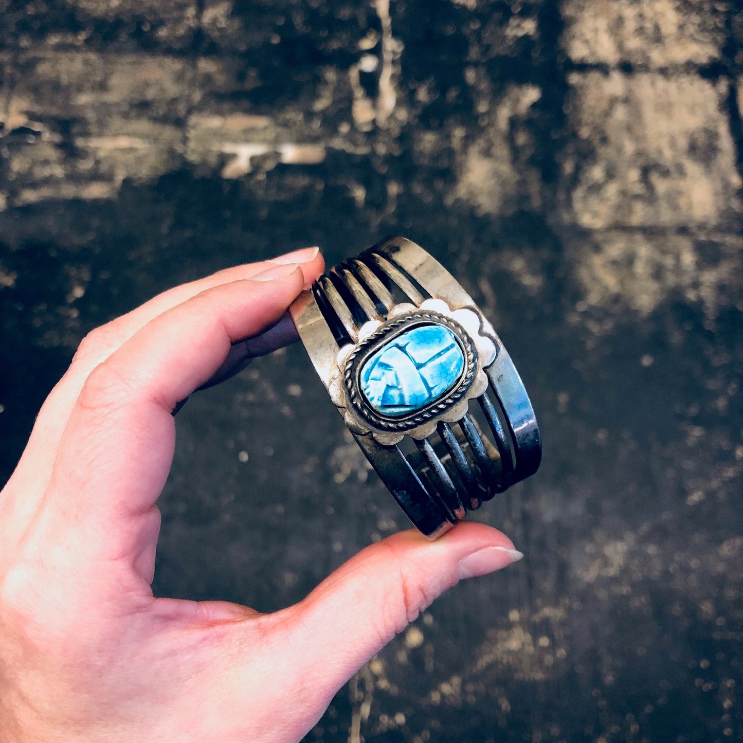 Vintage silver-toned wide cuff bracelet with large blue scarab cabochon held in hand with grungy weathered wooden background