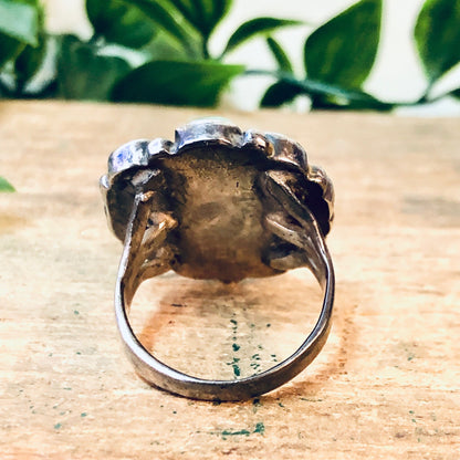 Vintage silver statement ring with large green turquoise stone in bohemian southwestern style, on rustic wooden surface with greenery in background.