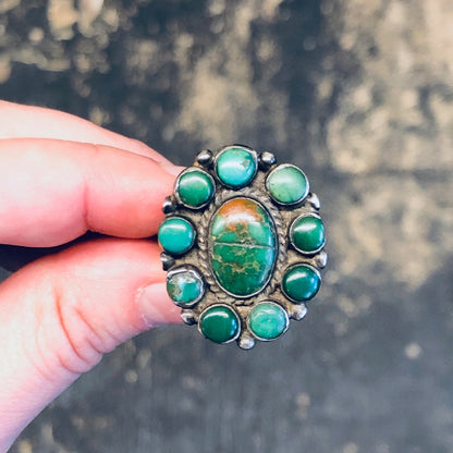 Large vintage silver ring featuring a central green turquoise cabochon surrounded by smaller turquoise stones, held in a person's hand against a textured dark gray background