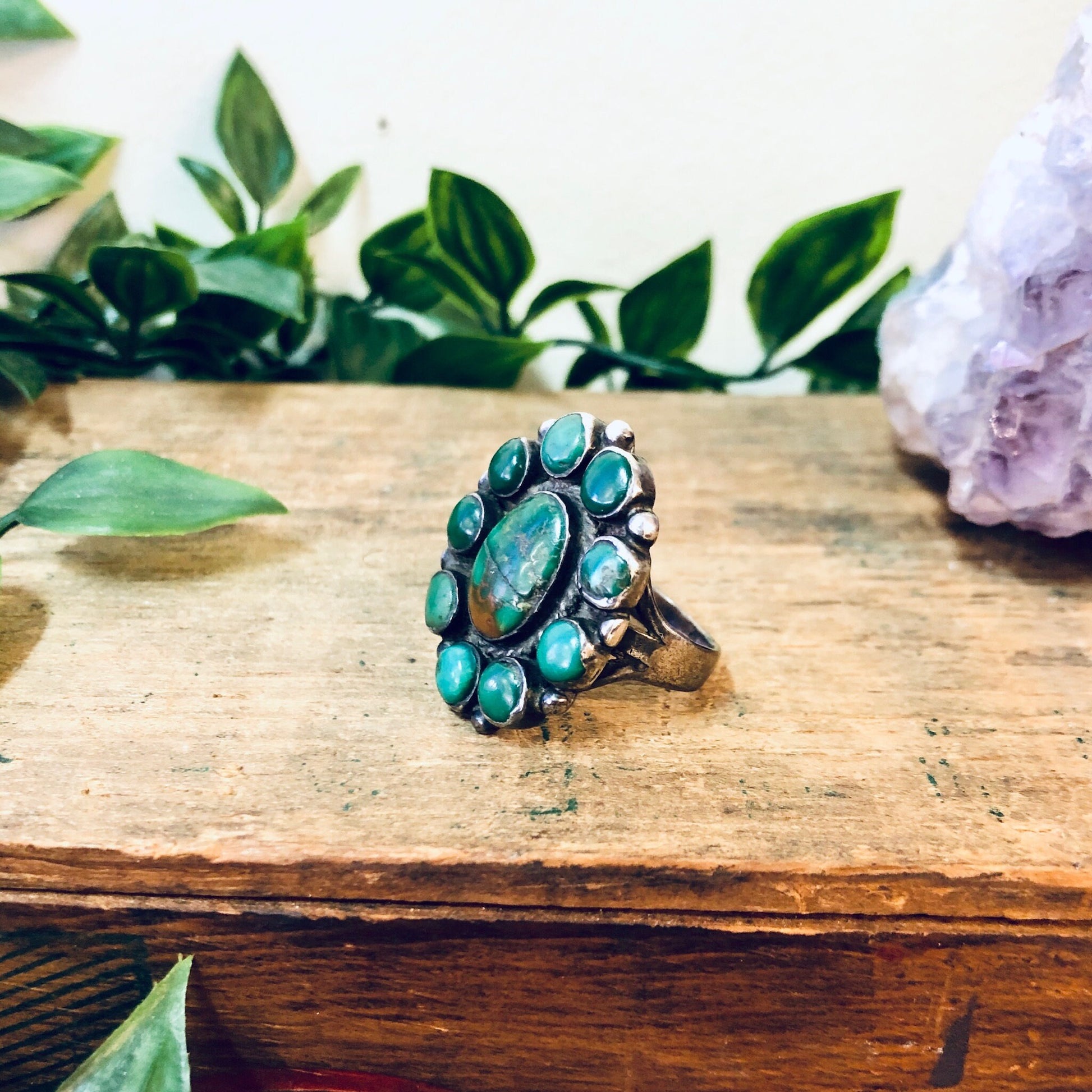 Large vintage silver ring featuring a cluster of green turquoise stones in a bohemian, Southwestern style statement piece, displayed on a wooden surface with plants in the background.