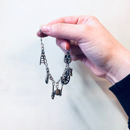 A hand holding a vintage-style silver charm bracelet with small hanging charms including scissors, against a plain background.