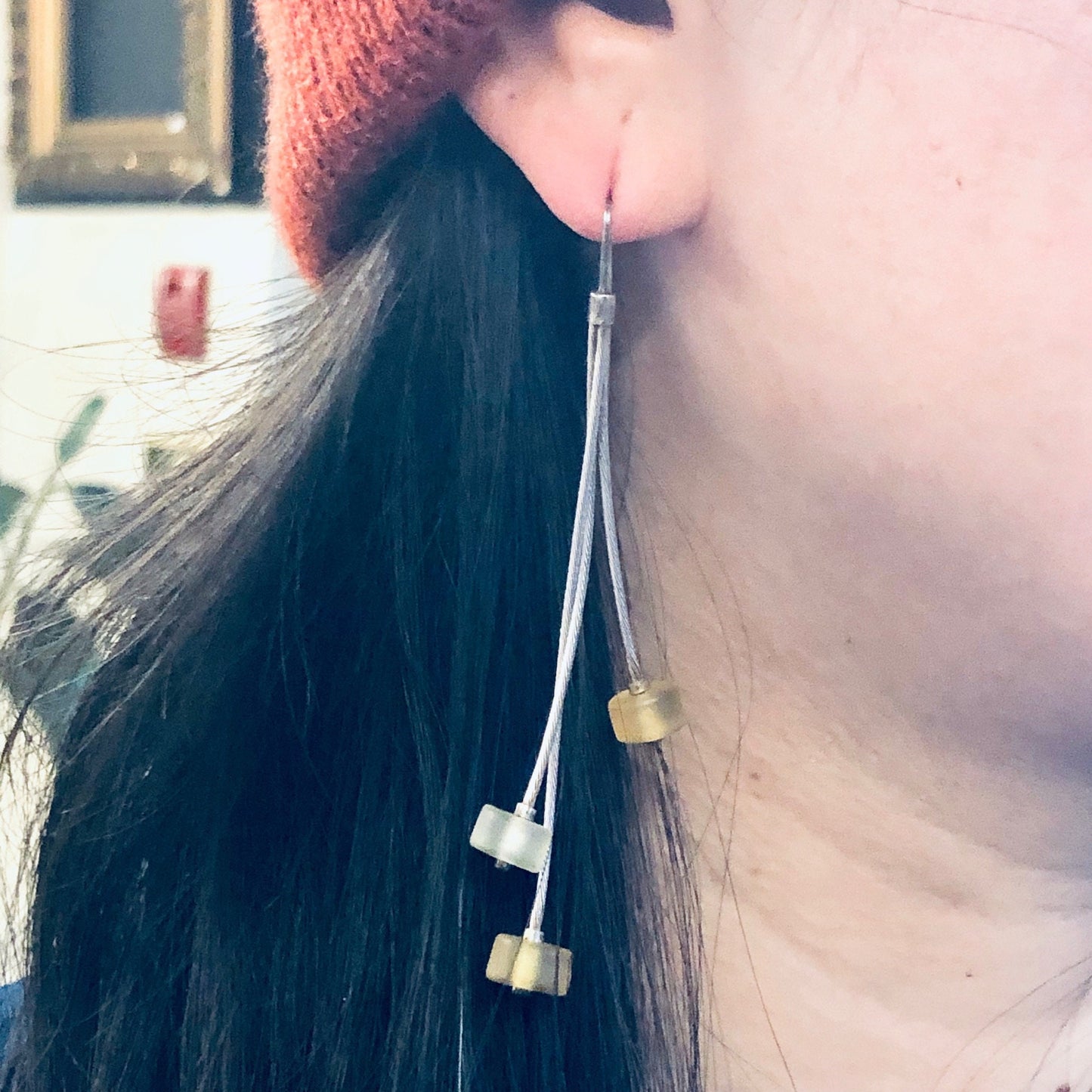 Close-up view of a person wearing long, dangling yellow and silver earrings alongside their dark hair, red knit hat and black top.