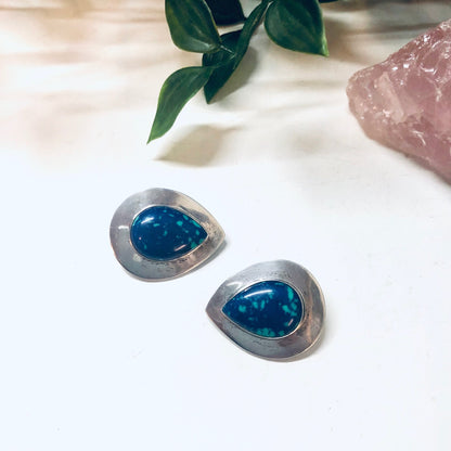 Vintage silver clip-on earrings with teardrop-shaped azurite malachite stones in shades of deep blue and green, displayed with rose quartz crystal and greenery on white background.