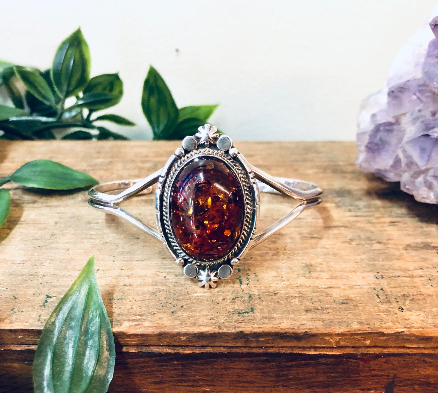 Vintage amber gemstone cuff bracelet in silver setting on wooden surface with green leaves and purple crystal.