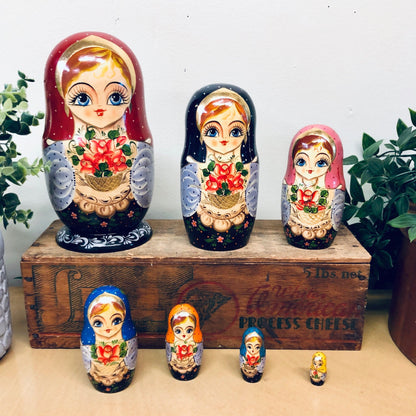 A set of seven vintage Russian nesting dolls, known as Matryoshka dolls, featuring hand-painted floral designs and adorable facial expressions. The wooden dolls are displayed on a wooden crate and surrounded by potted plants.