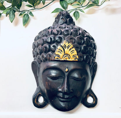 Vintage carved wooden Buddha mask with dark brown finish and gold accents, used as decorative wall hanging. Features serene facial expression and intricate detailing on headdress resembling leaves or petals. Asian art style home decor piece.