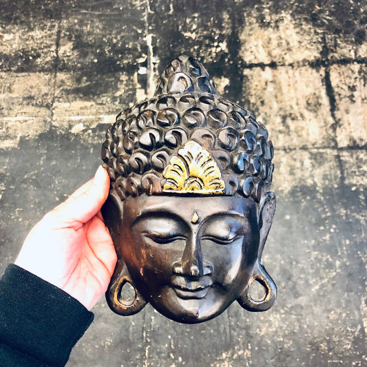 Carved wooden Buddha head mask with dark brown finish and gold accents, held in hand against weathered concrete wall background