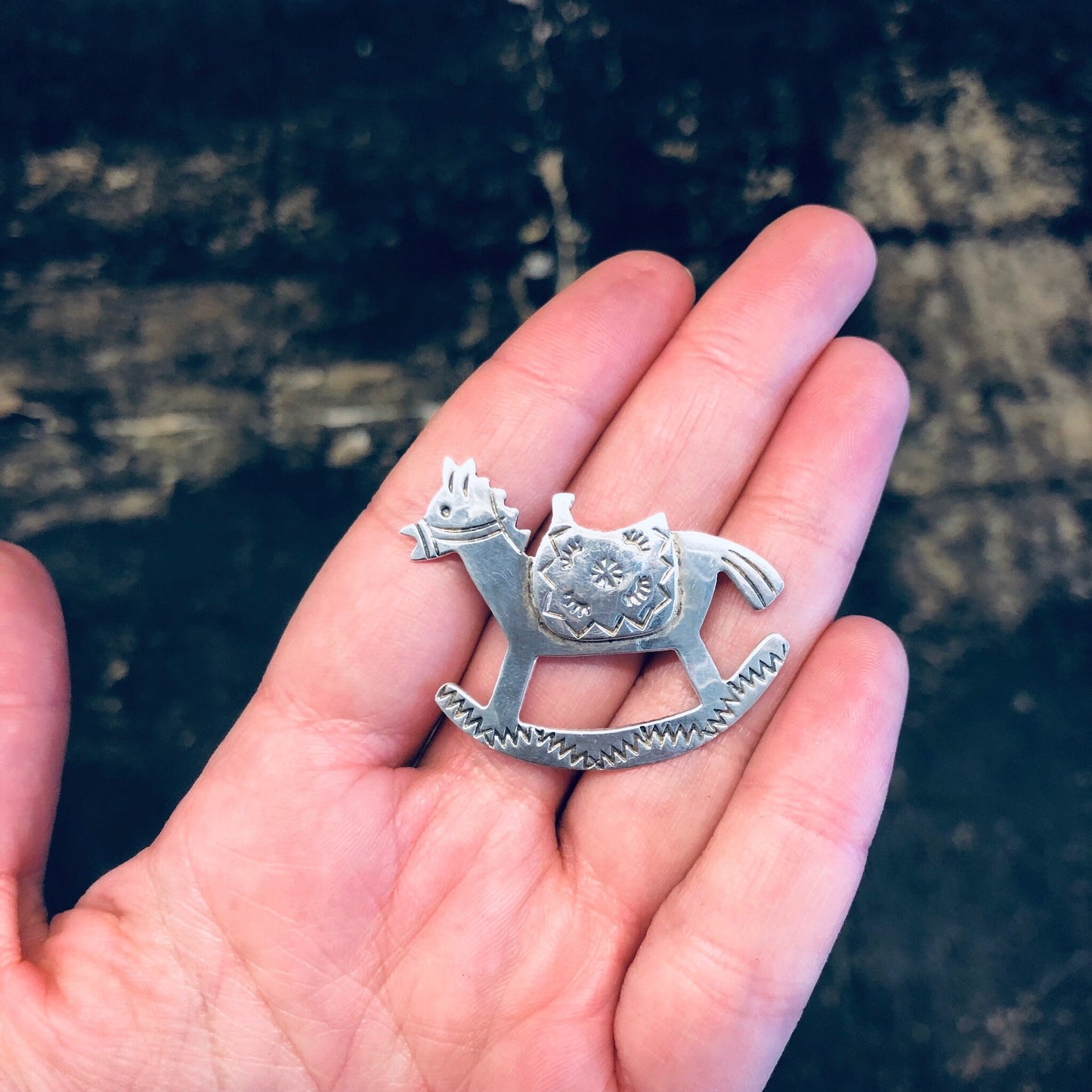 Vintage silver rocking horse brooch pin, made in Mexico, held in palm of hand against dark stone background