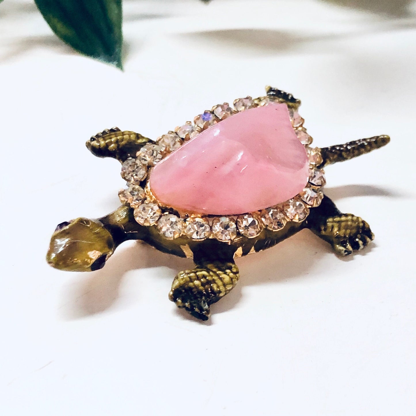 Vintage gold-toned turtle pin brooch with pink gemstone shell, clear rhinestones, and decorative feet, photographed on a white fabric background.