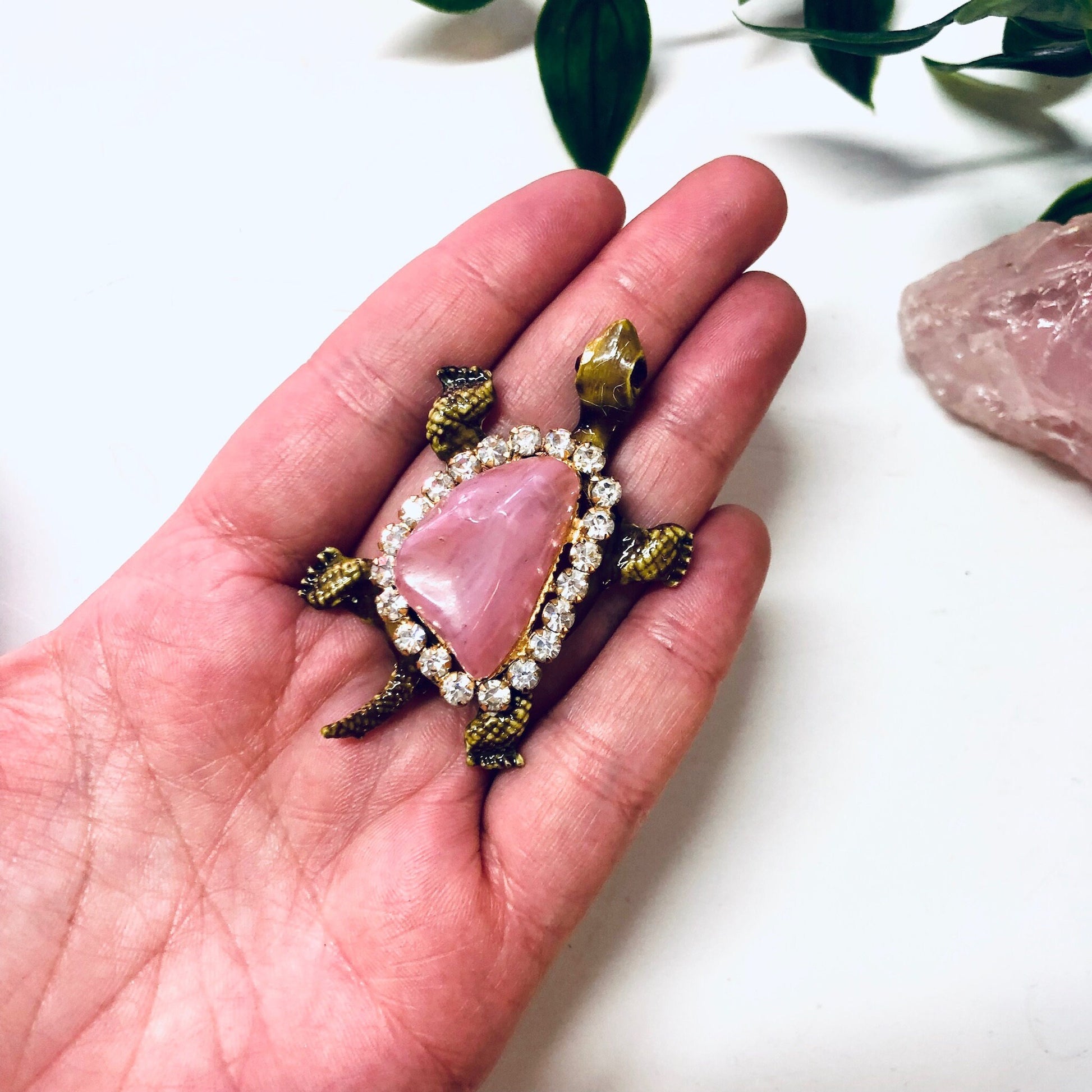 Vintage gold-toned turtle brooch pin with pink gemstone shell and rhinestone accents, held in an open palm against a white background with leaves and rose quartz crystal.