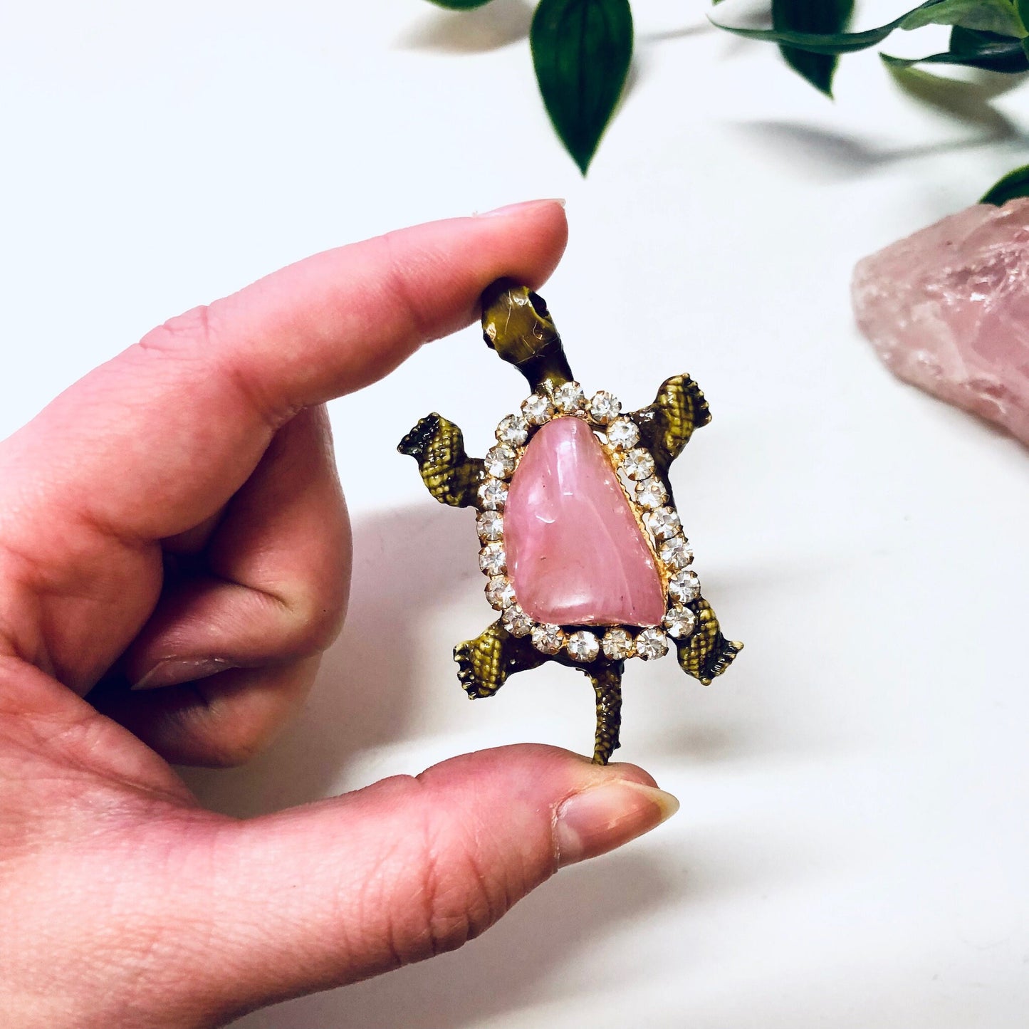 Vintage gold-toned turtle brooch with pink gemstone shell and rhinestone accents held in hand, with rose quartz crystal in background