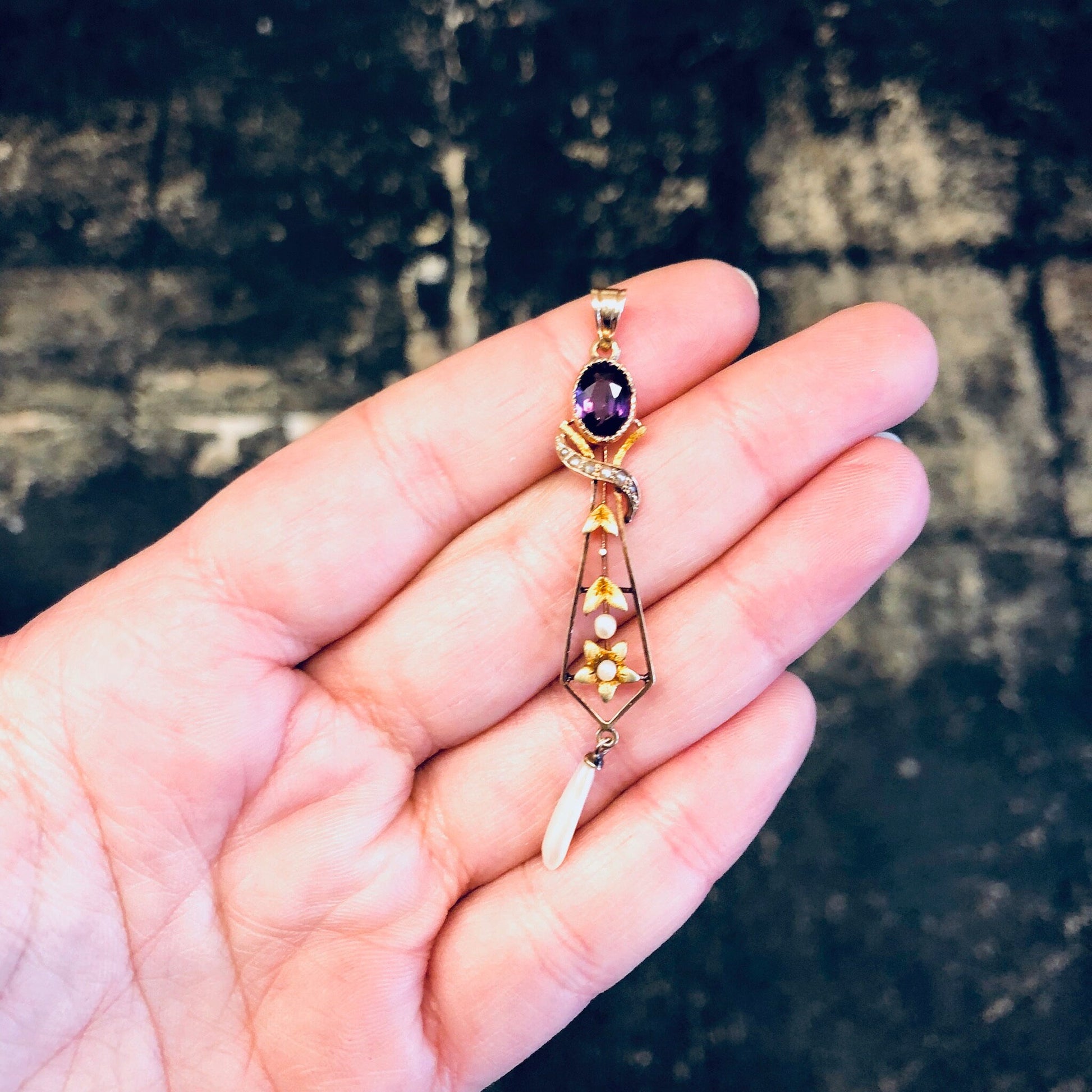 Vintage 14K yellow gold pendant necklace with amethyst stone and pearl accents in a floral design, held in palm of hand against weathered stone background.