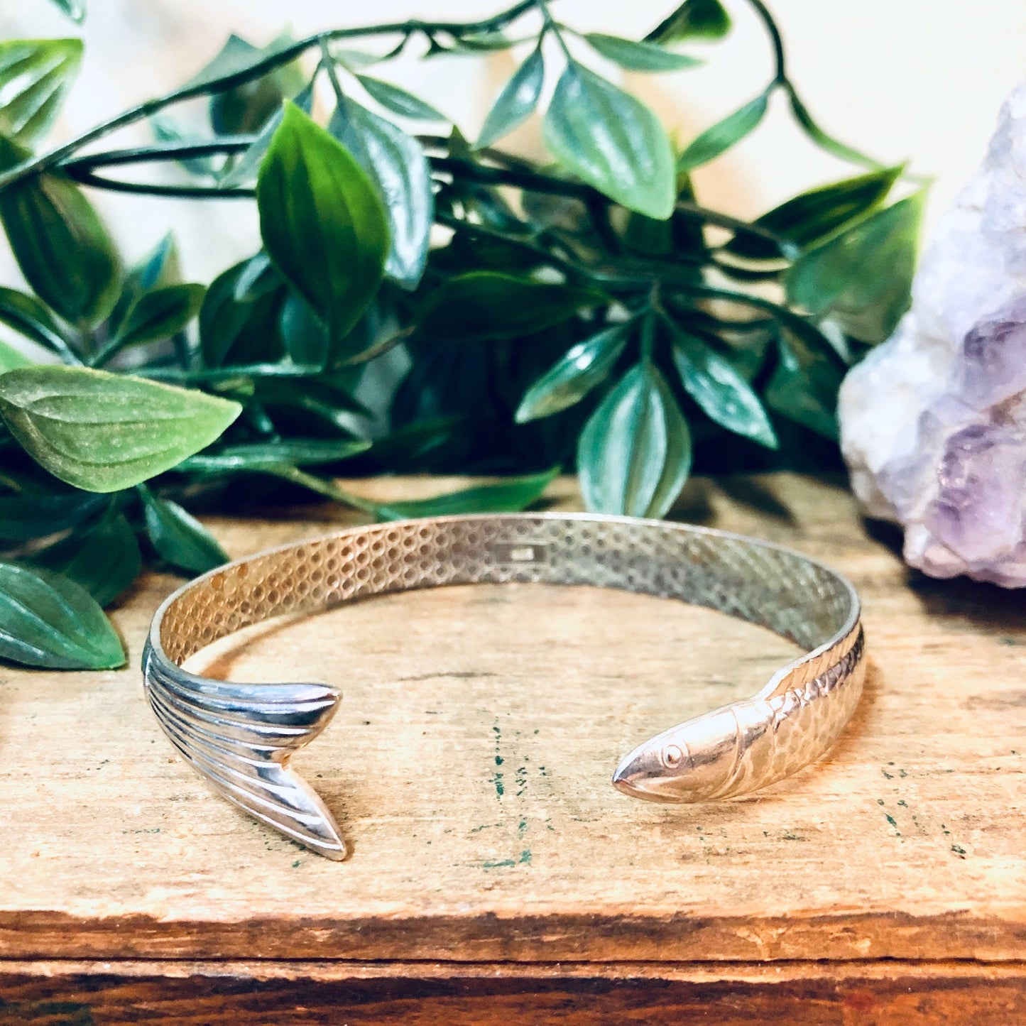 Silver fish bangle bracelet placed on wooden surface with green foliage and crystals in background, vintage aquatic animal jewelry cuff