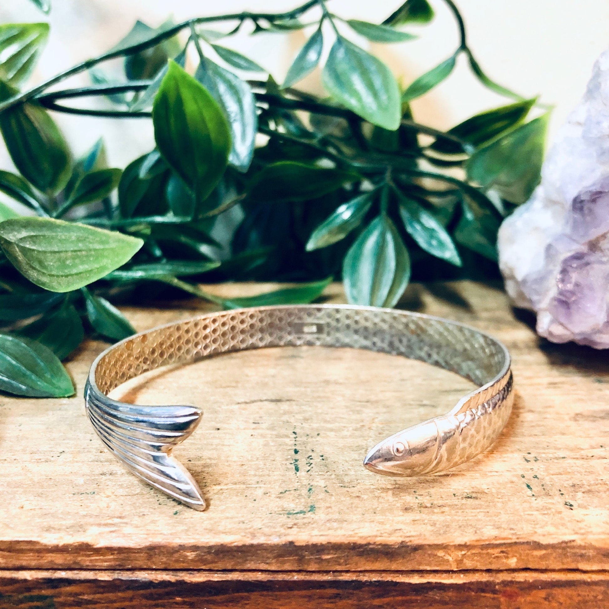 Silver fish bangle bracelet placed on wooden surface with green foliage and crystals in background, vintage aquatic animal jewelry cuff