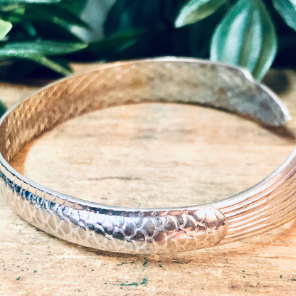 Vintage silver fish bangle cuff bracelet with hammered texture on weathered wooden surface with green foliage in background.