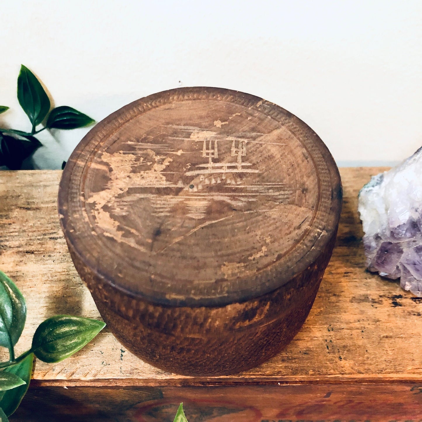 Round wooden trinket box with carved mountain landscape design, displayed on rustic wooden surface with green leaves and amethyst crystal.