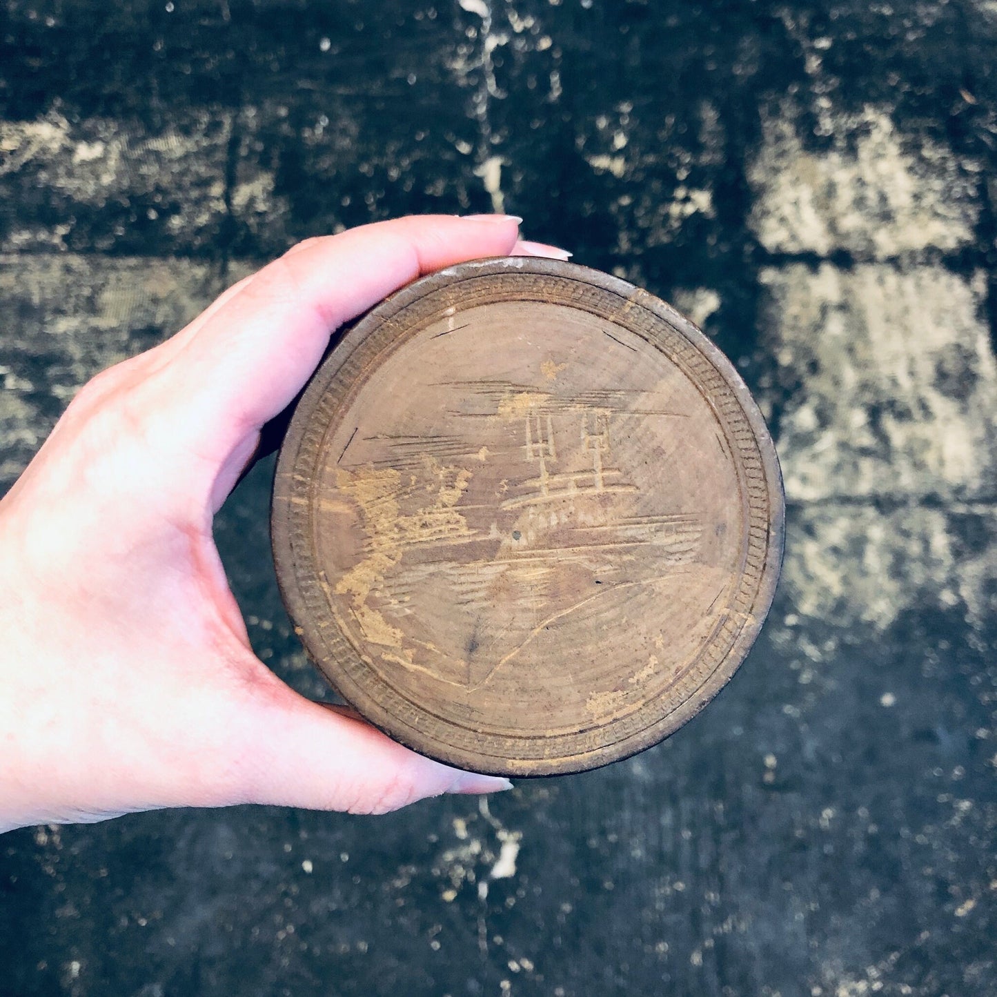 Hand holding a vintage, round wooden trinket box with intricate carvings, against a grungy concrete background.