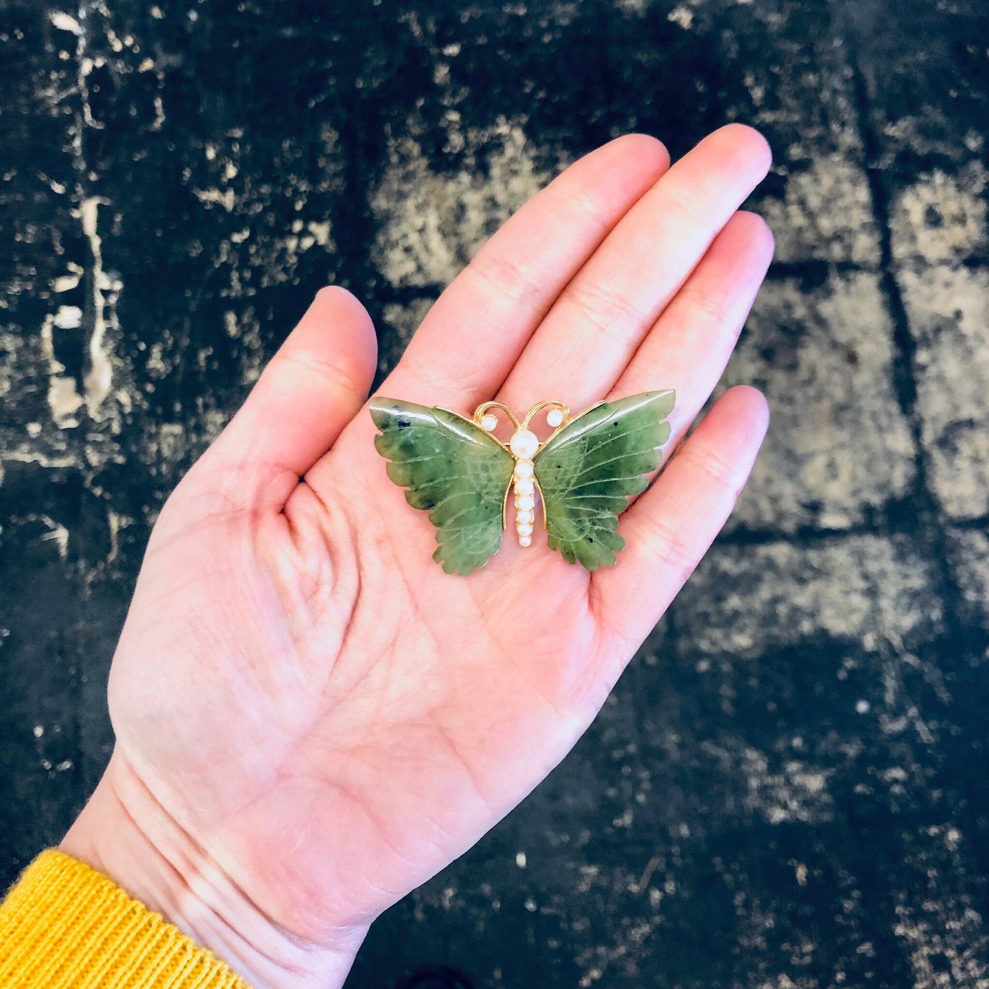 Hand holding a vintage gold-toned butterfly brooch with green jade wings and faux pearls against a grungy stone surface