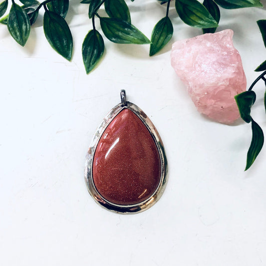 Vintage large druzy quartz pendant with a polished sparkly stone in a metal setting displayed on a white surface with green leaves and a pink crystal in the background for statement jewelry.