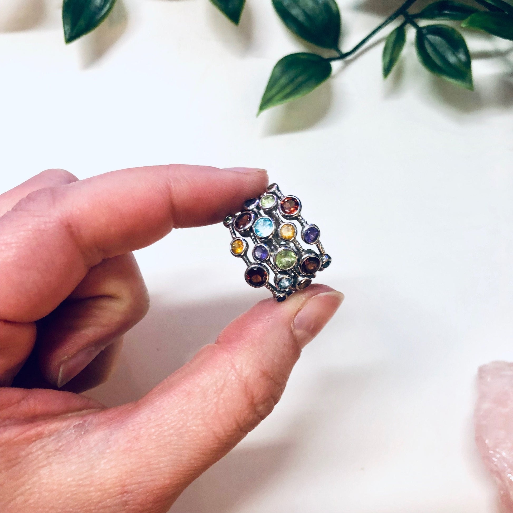 Vintage silver multi-band ring with colorful gemstones in purple, blue, red, green and yellow hues, held between fingers with leaves in soft focus background.