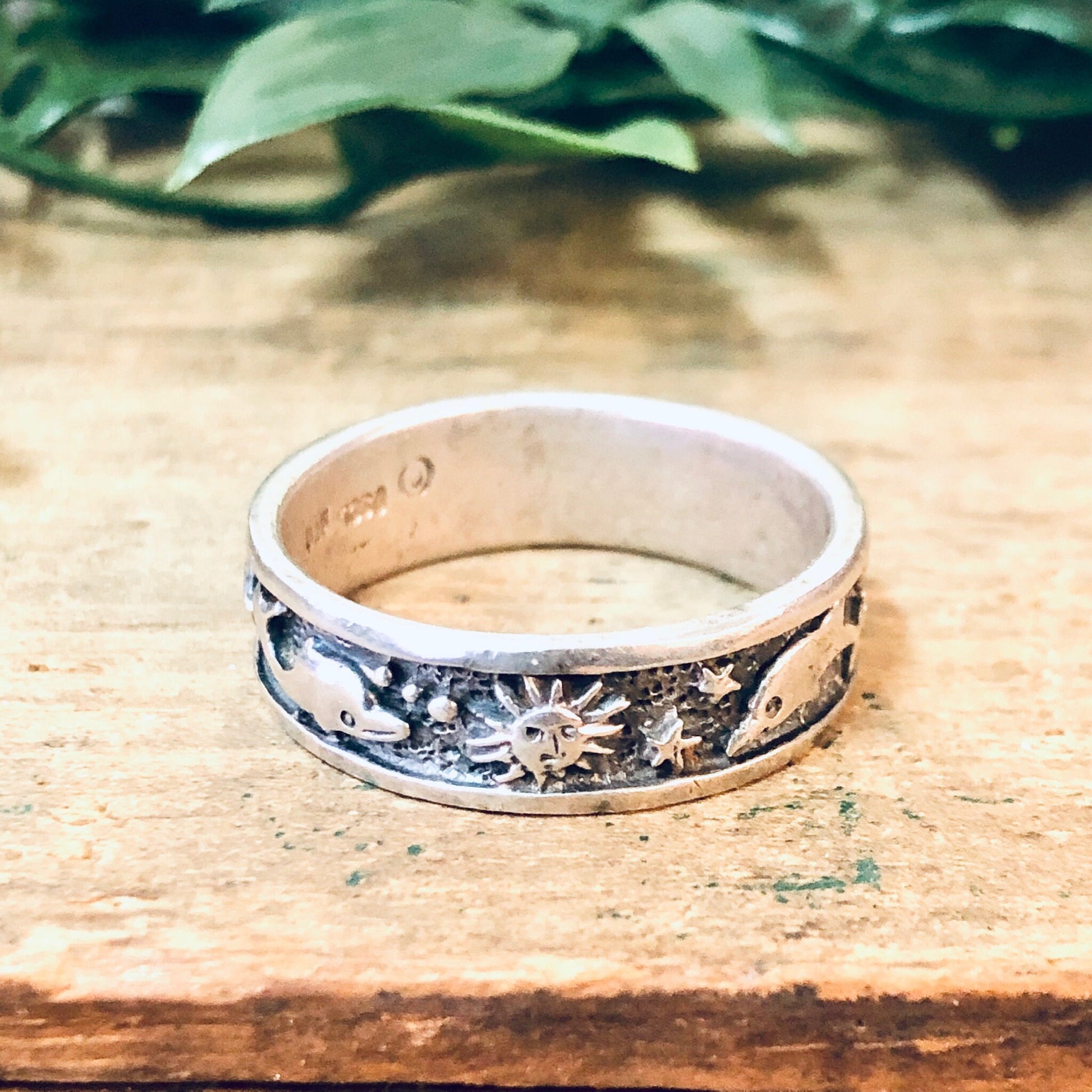 Vintage silver ring with aquatic design featuring dolphins, sun and ocean motifs, worn on weathered wooden surface with leaves in background.