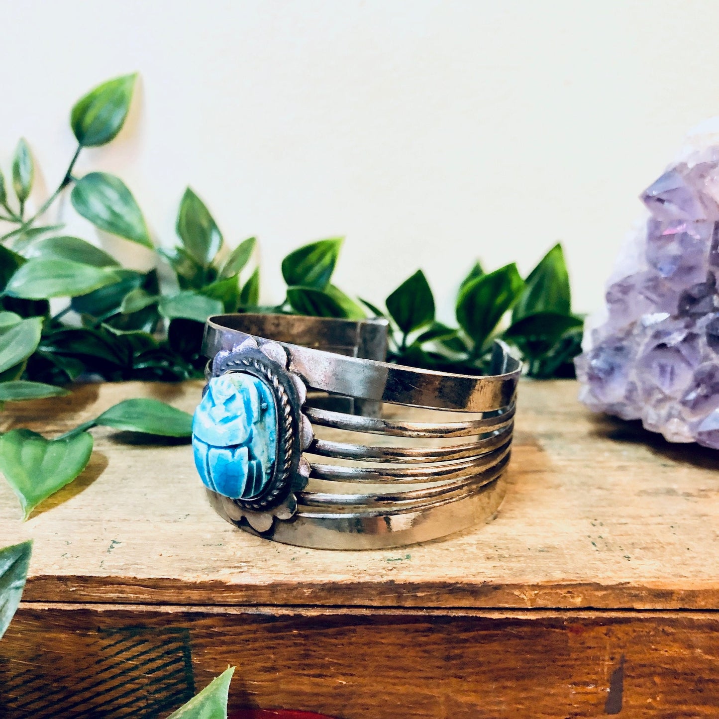 Vintage silver-toned cuff bracelet with large blue scarab gemstone, surrounded by green leaves, on a wooden surface next to an amethyst crystal.