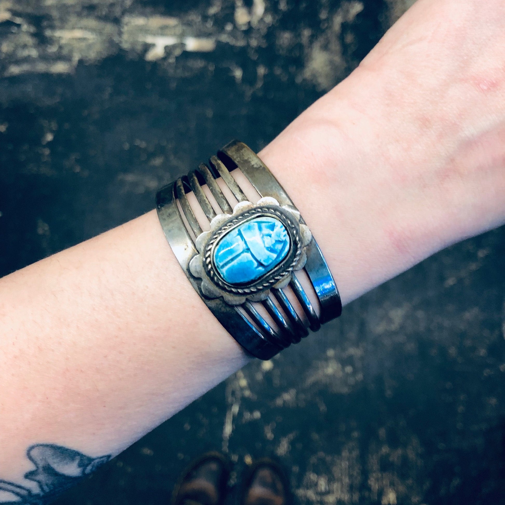 Vintage silver-toned cuff bracelet with large blue scarab beetle design, worn on a wrist against a dark distressed background.