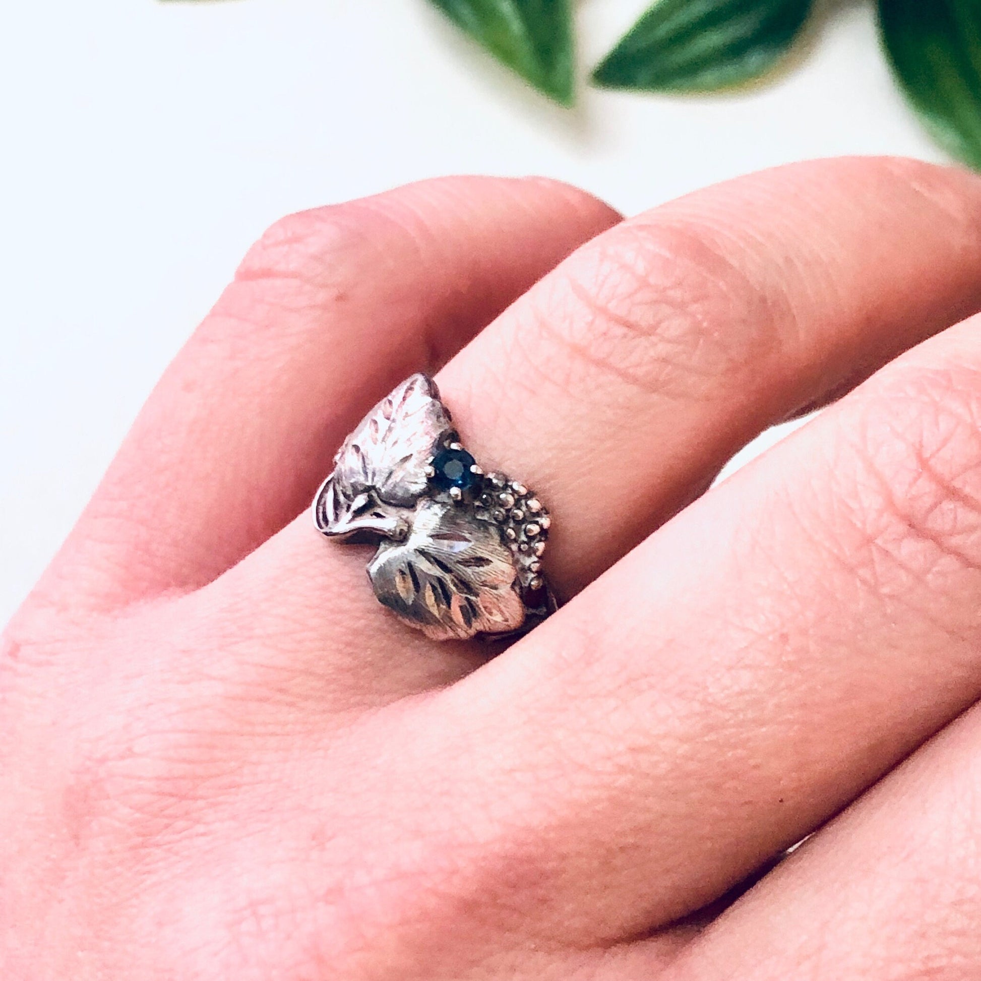 Vintage silver leaf ring with blue gemstone and etched design, worn on finger, close-up view with greenery in background - anniversary gift or birthstone jewelry in 925 silver.